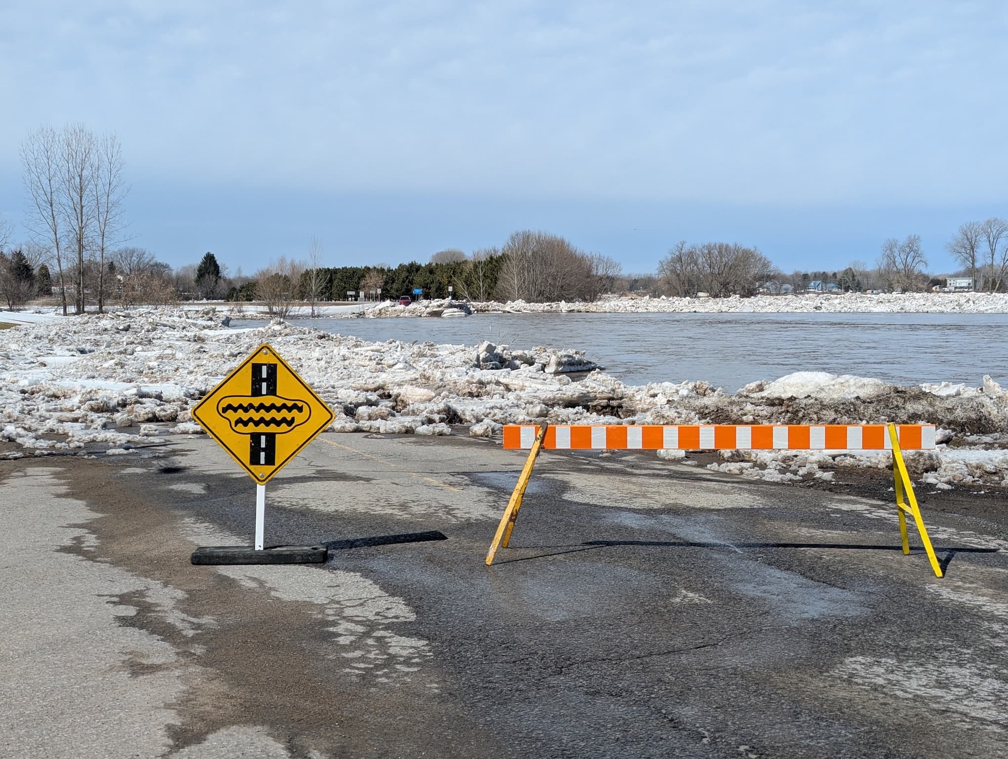 Flooded road with warning sign and barrier, surrounded by snow and ice.